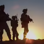 Squad of Three Fully Equipped and Armed Soldiers Standing on Hill in Desert Environment in Sunset Light.